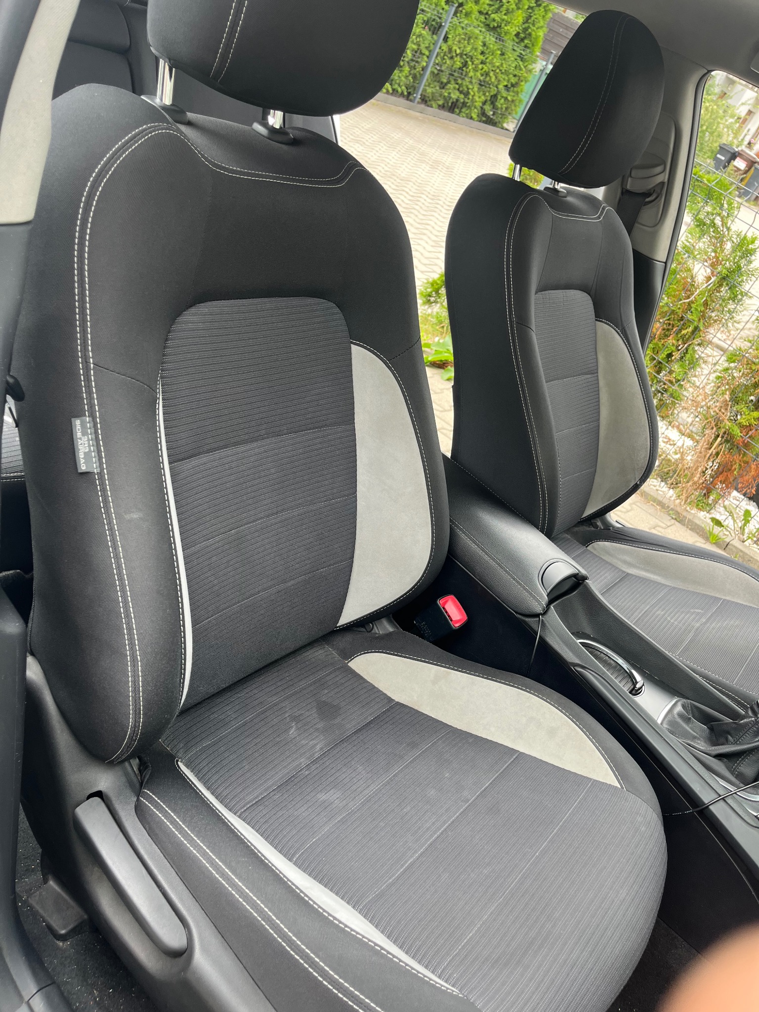 Dwa czarne fotele samochodowe z szarymi wstawkami i białymi szwami, widoczne z bliska, z zewnątrz auta w tle widać zieleń.