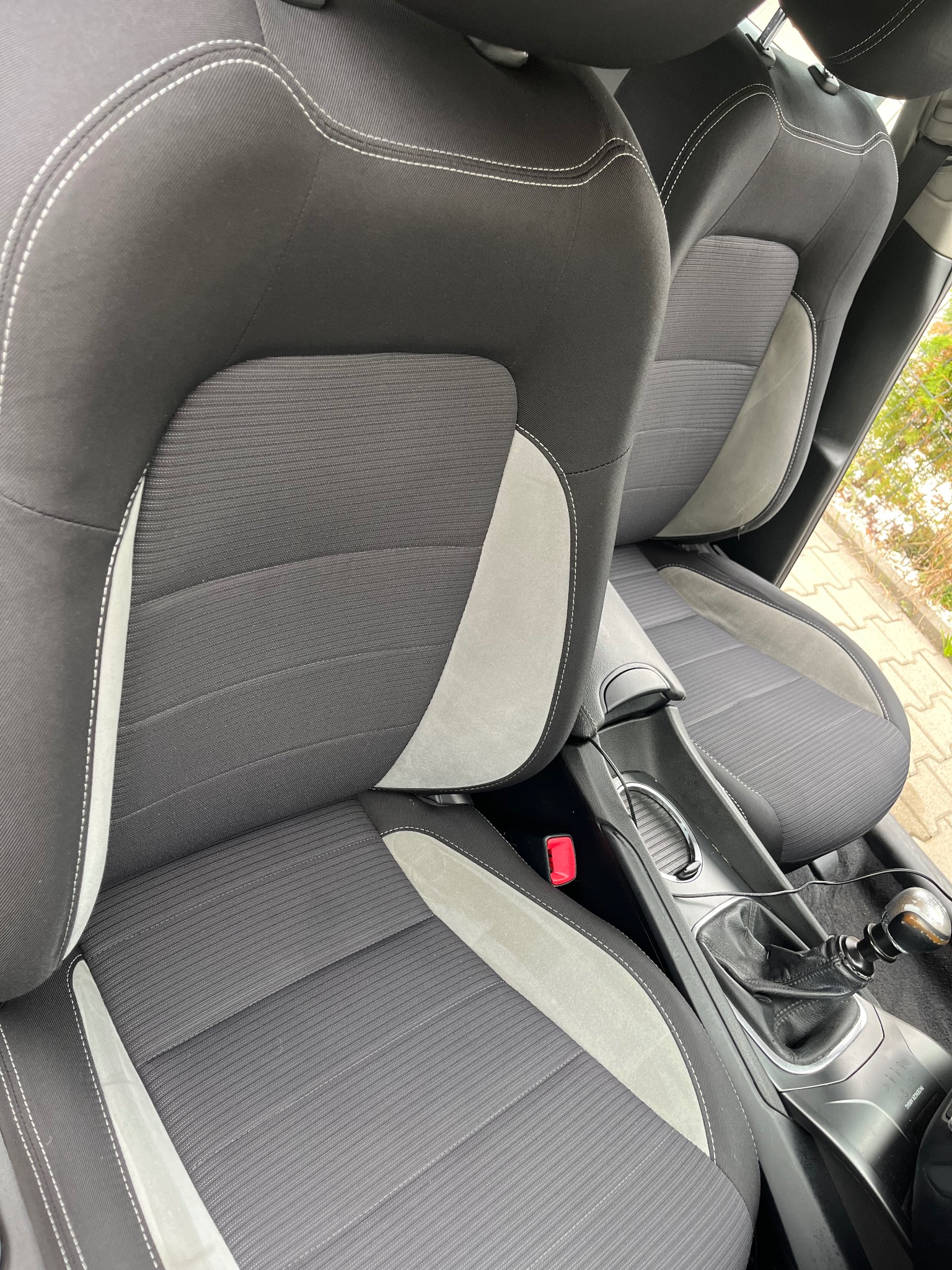 Wnętrze samochodu z widocznymi czarno-szarymi fotelami z materiału, szwami w kolorze ecru, konsolą środkową z dźwignią zmiany biegów i fragmentem pasa bezpieczeństwa.