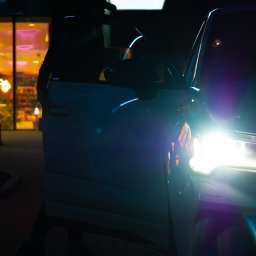 MANEE Group Mateusz Zacharow - Sylwetka osoby w czapce wsiadającej do białego samochodu w nocnym otoczeniu miejskim, z widocznymi światłami samochodu i neonami w tle.