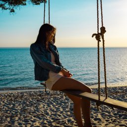 MANEE Group Mateusz Zacharow - Kobieta w dżinsowej kurtce siedzi na drewnianej huśtawce na plaży, spoglądając w stronę morza o zachodzie słońca.