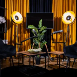 Studio podcastowe z dwoma krzesłami, mikrofonami i lampami studyjnymi na tle czarno-złotych zasłon.