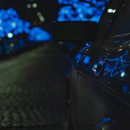 MANEE Group Mateusz Zacharow - Częściowy widok ciemnego samochodu zaparkowanego na ulicy w nocy, z rozmytymi niebieskimi światłami w tle.