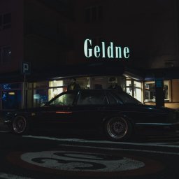 MANEE Group Mateusz Zacharow - Czarny, klasyczny samochód zaparkowany na ulicy nocą, z neonowym napisem 'Geldne' widocznym w tle nad budynkiem.