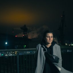 MANEE Group Mateusz Zacharow - Portret kobiety w szarym płaszczu i czarnej bluzie z kapturem, na tle nocnego krajobrazu z dźwigami portowymi i oświetleniem miejskim.