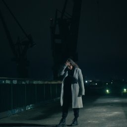 MANEE Group Mateusz Zacharow - Kobieta w płaszczu stoi na tle dźwigów portowych nocą, zakrywając twarz dłonią.