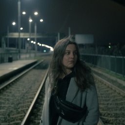 MANEE Group Mateusz Zacharow - Portret kobiety w szarym płaszczu i czarnej nerce stojącej na torach kolejowych nocą, oświetlonych przez lampy uliczne.