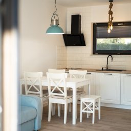 MANEE Group Mateusz Zacharow - Jasna kuchnia z białymi meblami, drewnianym blatem i czarnymi akcentami, oświetlona turkusową lampą wiszącą i lampą z linami, widziana zza rogu pokoju z błękitnym fotelem.