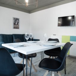 MANEE Group Mateusz Zacharow - Nowoczesne wnętrze jadalni z białym stołem nakrytym dla czterech osób, czarne krzesła, szara sofa i kolorowa komoda pod telewizorem.