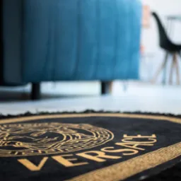 Okrągły, czarny dywan z wyraźnym złotym wzorem Versace, częściowo widoczny niebieski narożnik sofy i czarne krzesło w tle.