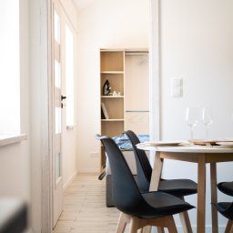 MANEE Group Mateusz Zacharow - Jasne wnętrze jadalni z okrągłym stołem, czarnymi krzesłami i widokiem na garderobę w tle.