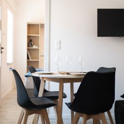 MANEE Group Mateusz Zacharow - Jasne wnętrze mieszkania z okrągłym stołem nakrytym dla trzech osób, czarne krzesła z drewnianymi nogami, telewizor na ścianie, widok na sypialnię w tle.