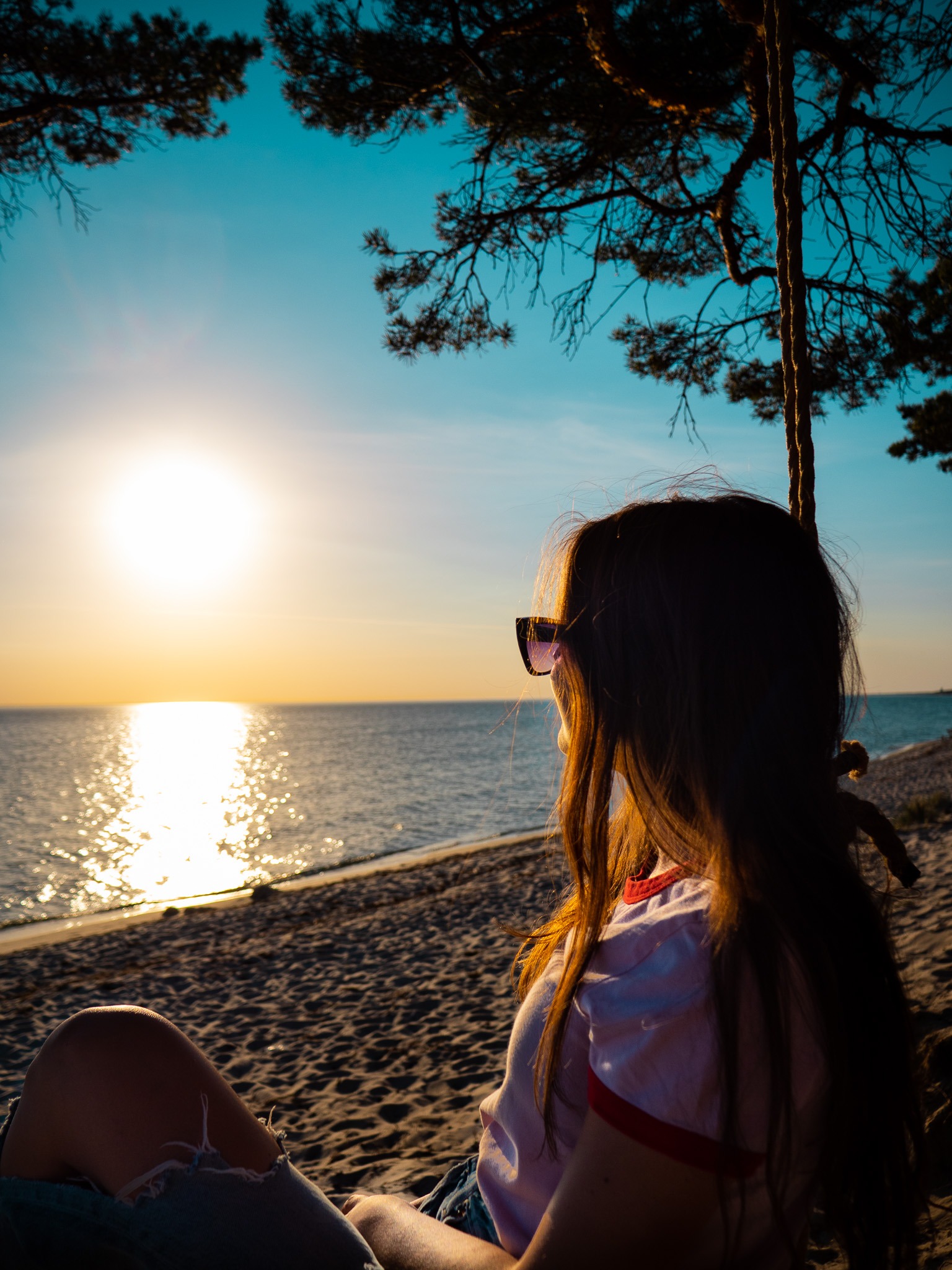 Dziewczyna w okularach przeciwsłonecznych siedzi na huśtawce na plaży, obserwując zachód słońca nad morzem.