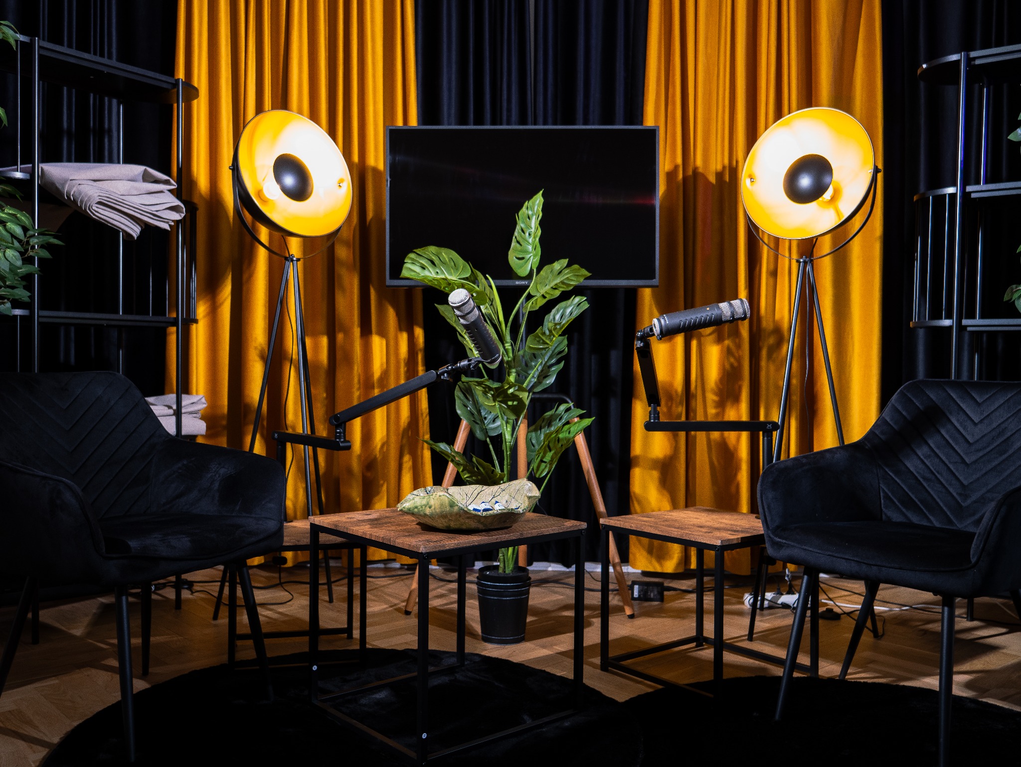 Studio podcastowe z dwoma krzesłami, mikrofonami i lampami studyjnymi na tle czarno-złotych zasłon.