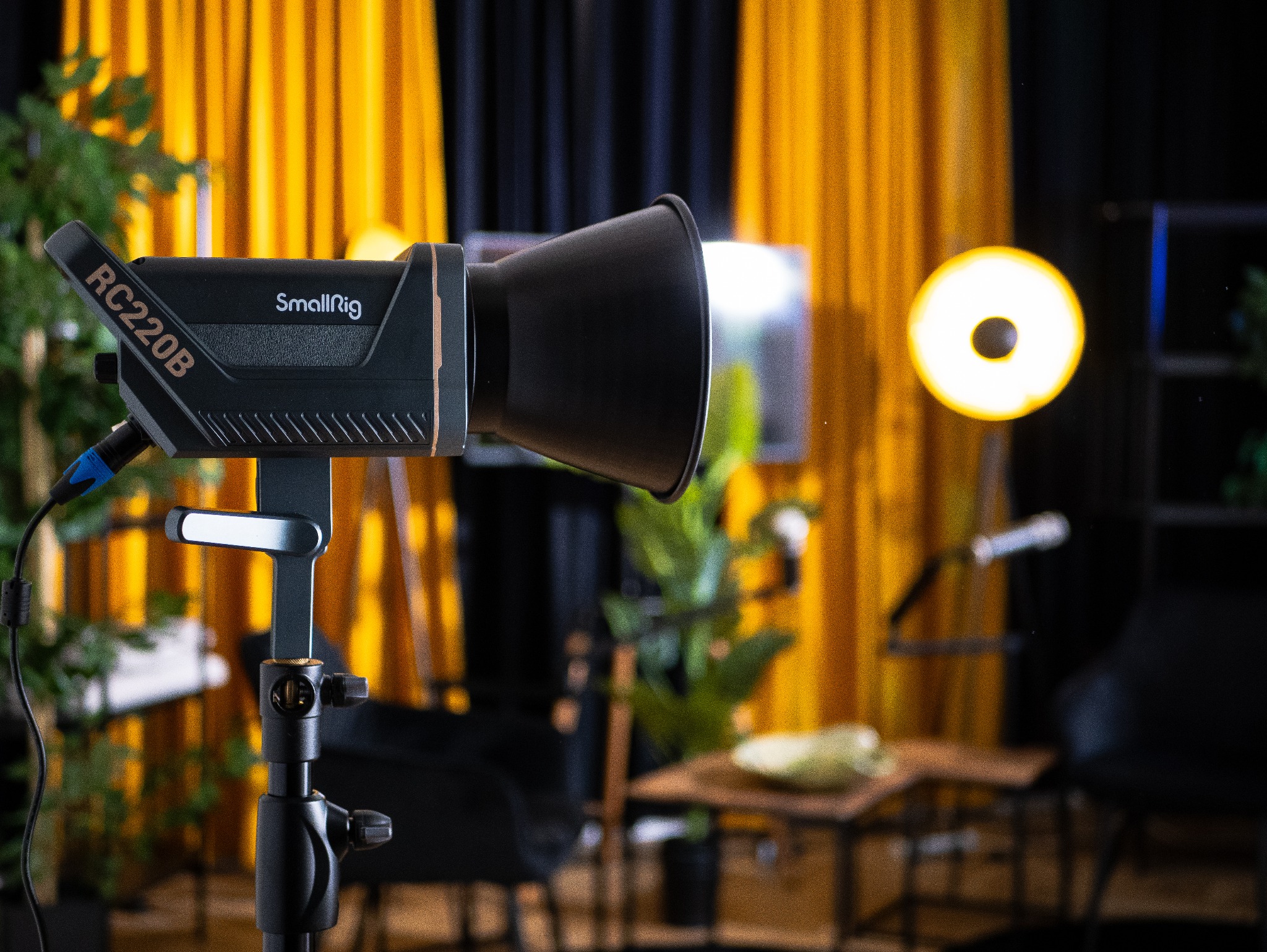 Lampa studyjna SmallRig RC220B na statywie, z widocznym kablem zasilającym i światłem, w tle żółte i czarne zasłony oraz rozmyte elementy wyposażenia studia.