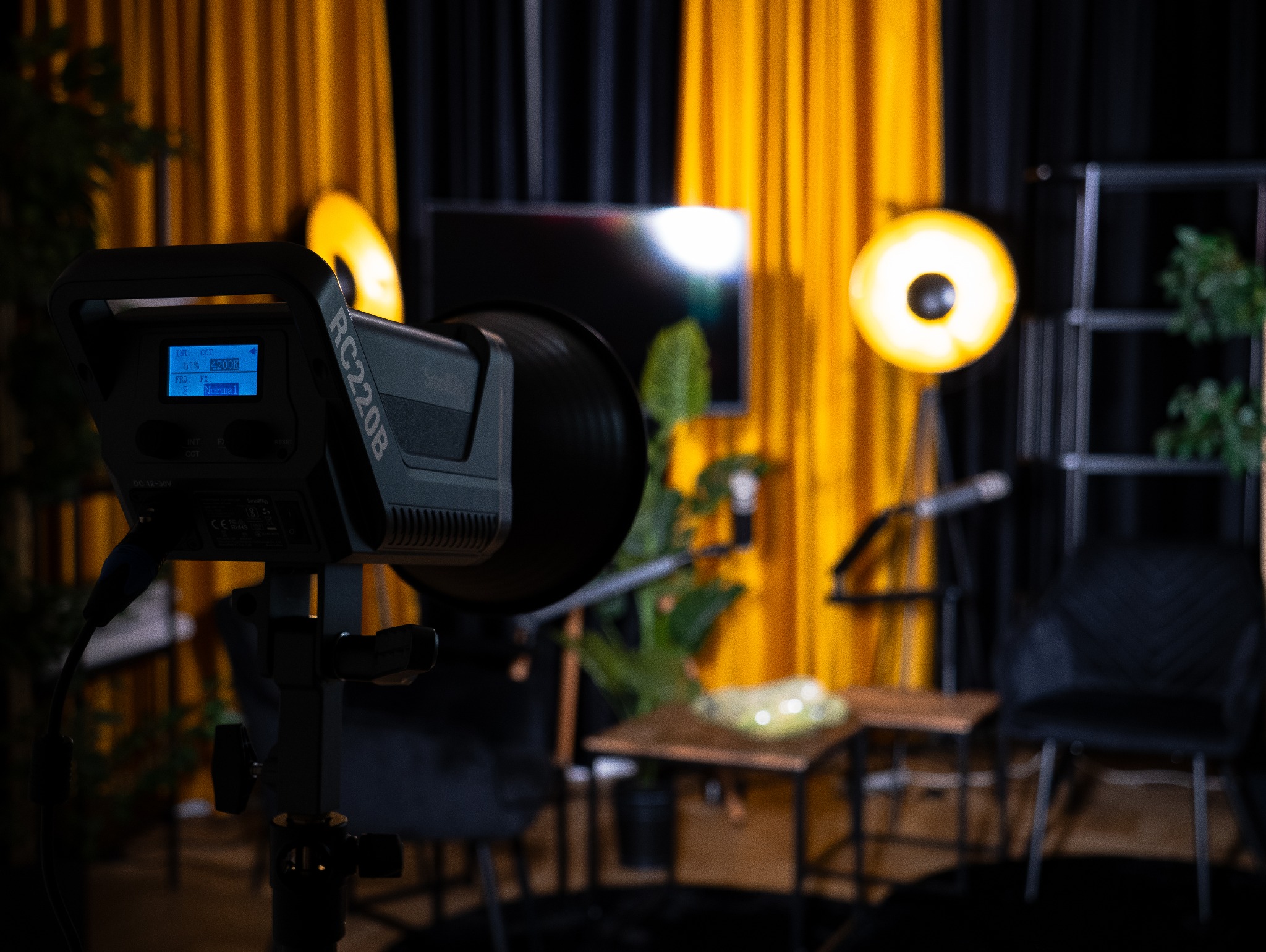 Lampa studyjna RC220B z wyświetlaczem ustawień, ustawiona w studio w Połczynie z żółtymi zasłonami w tle, obok widoczna roślina doniczkowa i mikrofon.
