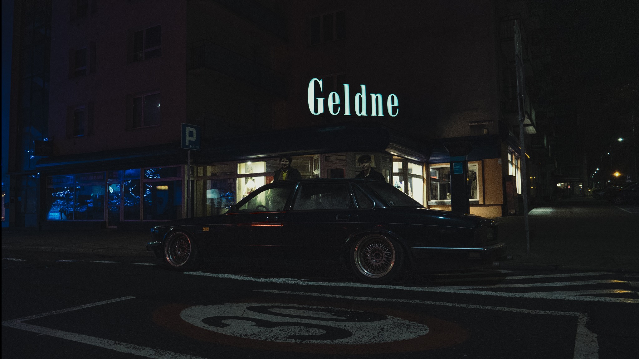 Czarny, klasyczny samochód zaparkowany na ulicy nocą, z neonowym napisem 'Geldne' widocznym w tle nad budynkiem.