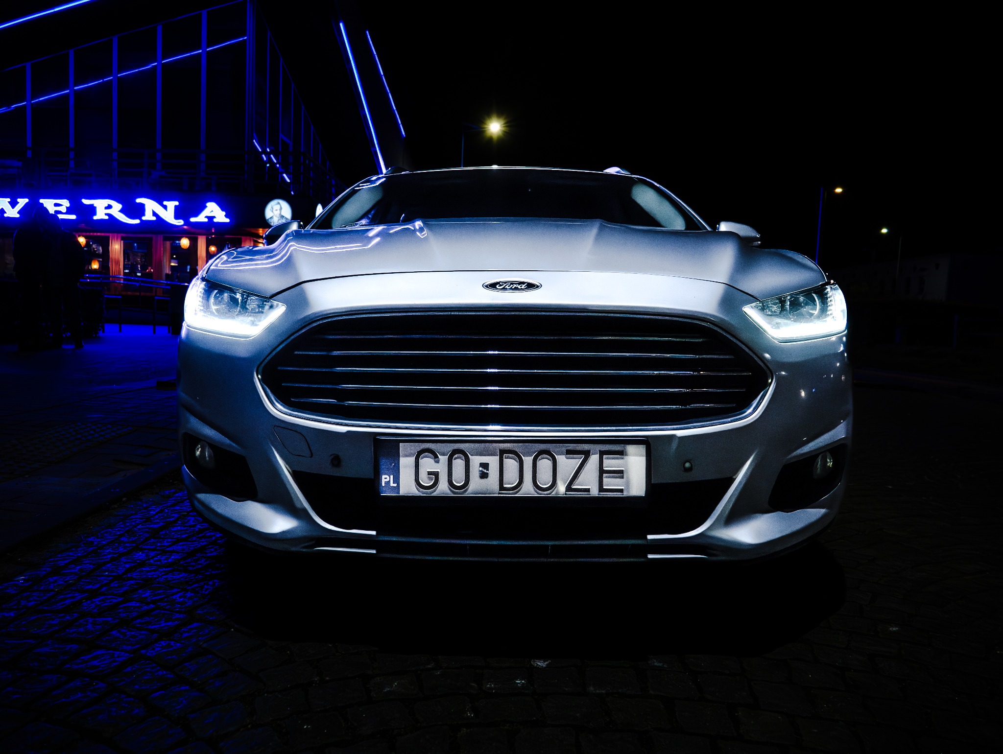 Przód srebrnego samochodu Ford z włączonymi światłami, tablica rejestracyjna 'GO-DOZE', na tle nocnego krajobrazu z budynkiem oświetlonym na niebiesko.