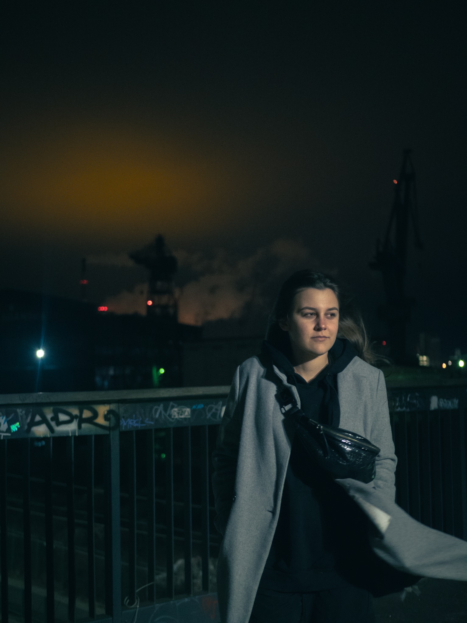 Portret kobiety w szarym płaszczu i czarnej bluzie z kapturem, na tle nocnego krajobrazu z dźwigami portowymi i oświetleniem miejskim.