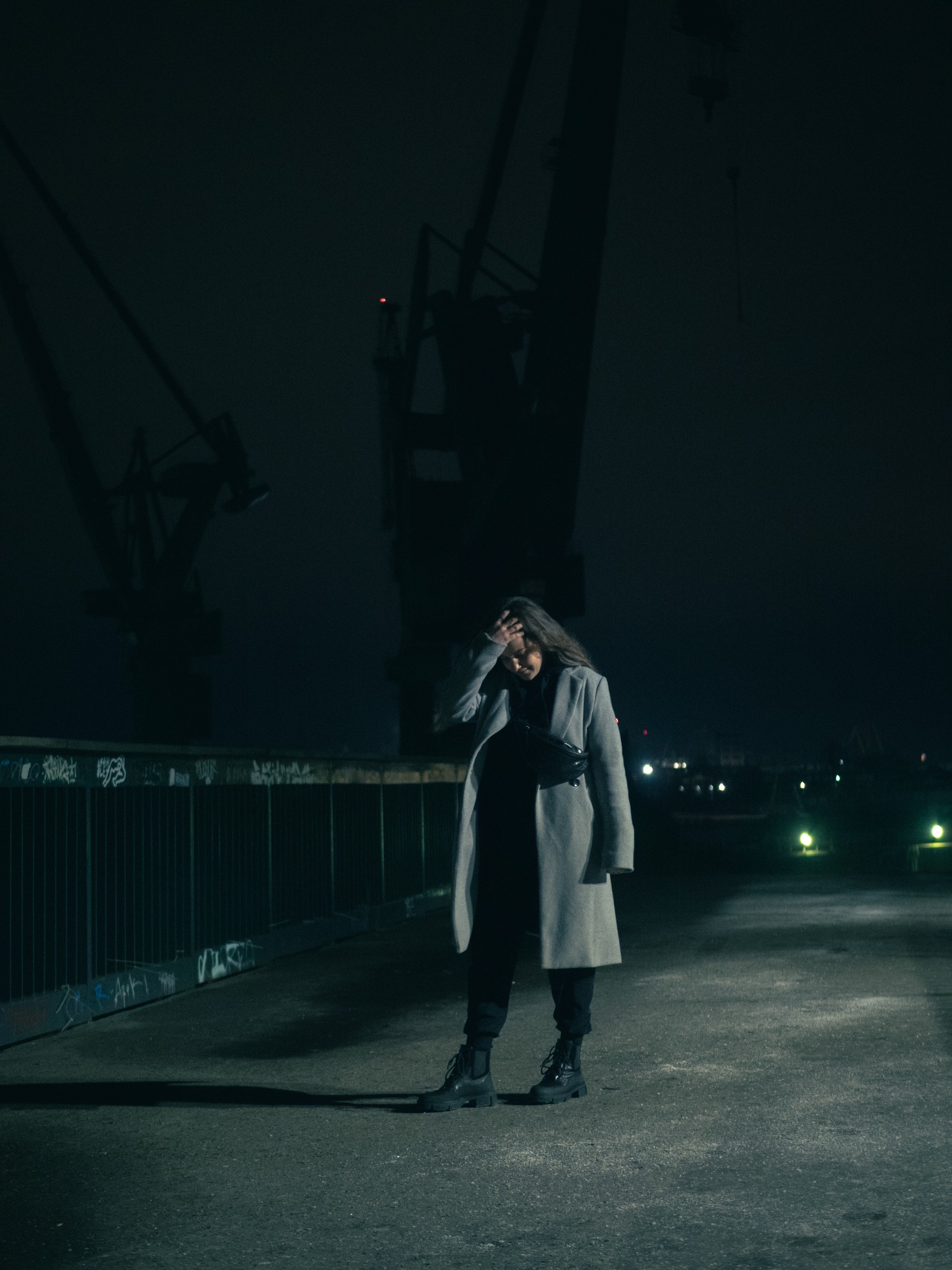 Kobieta w płaszczu stoi na tle dźwigów portowych nocą, zakrywając twarz dłonią.