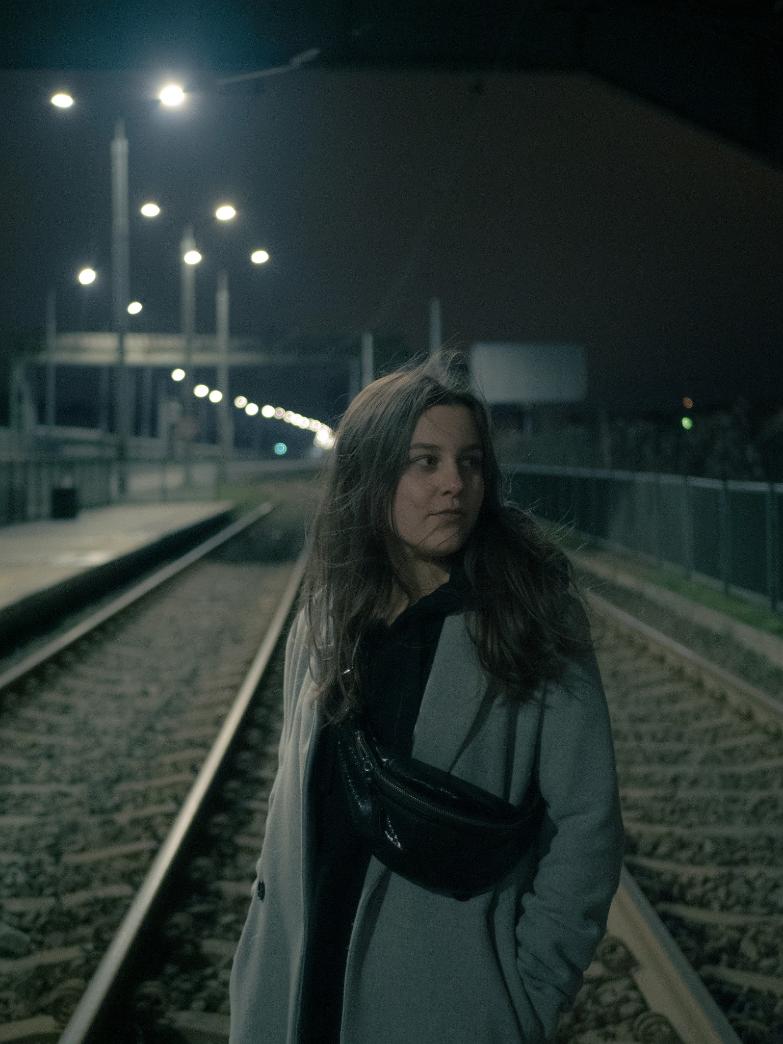 Portret kobiety w szarym płaszczu i czarnej nerce stojącej na torach kolejowych nocą, oświetlonych przez lampy uliczne.