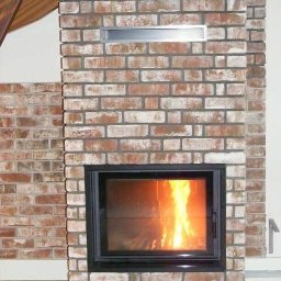 Kominkiportalowe - Murowany kominek z cegły z płomieniami widocznymi przez szybę w Buczkowicach, z metalową wnęką powyżej paleniska.