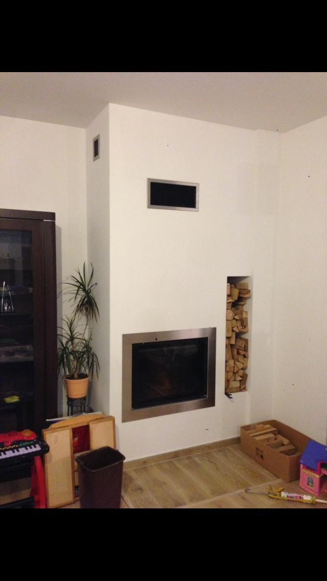 Nowoczesny kominek z metalową ramą w białej obudowie, z wnęką na drewno po prawej stronie i kratką wentylacyjną nad paleniskiem, otoczenie w trakcie prac wykończeniowych.