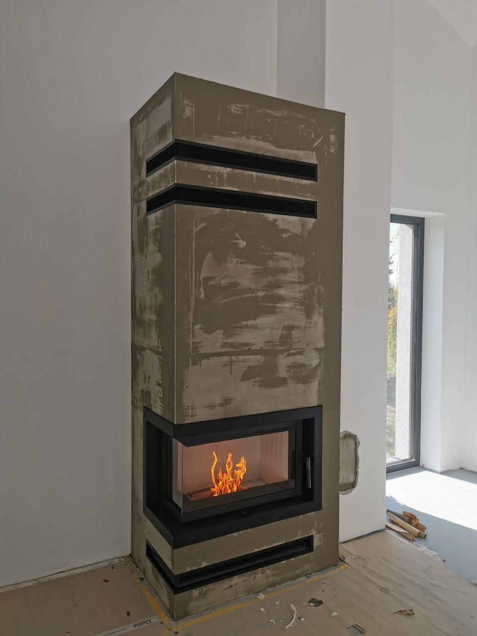 Nowoczesny kominek narożny w trakcie budowy, z widocznym paleniskiem i surową obudową w odcieniach szarości, czarne elementy dekoracyjne, niedokończone wykończenie ściany obok okna.