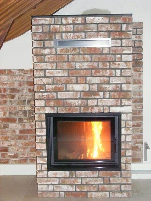 Murowany kominek z cegły z płomieniami widocznymi przez szybę w Buczkowicach, z metalową wnęką powyżej paleniska.