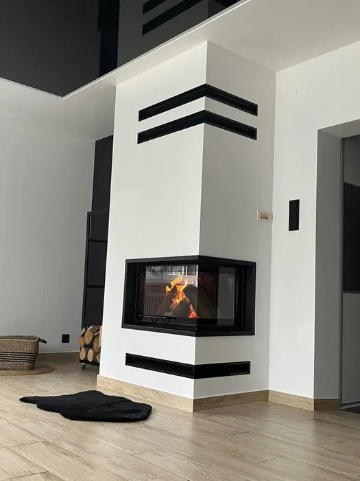Nowoczesny kominek narożny z czarną obudową i dekoracyjnymi poziomymi pasami, płomienie widoczne przez szybę, na drewnianej podłodze czarny dywanik.
