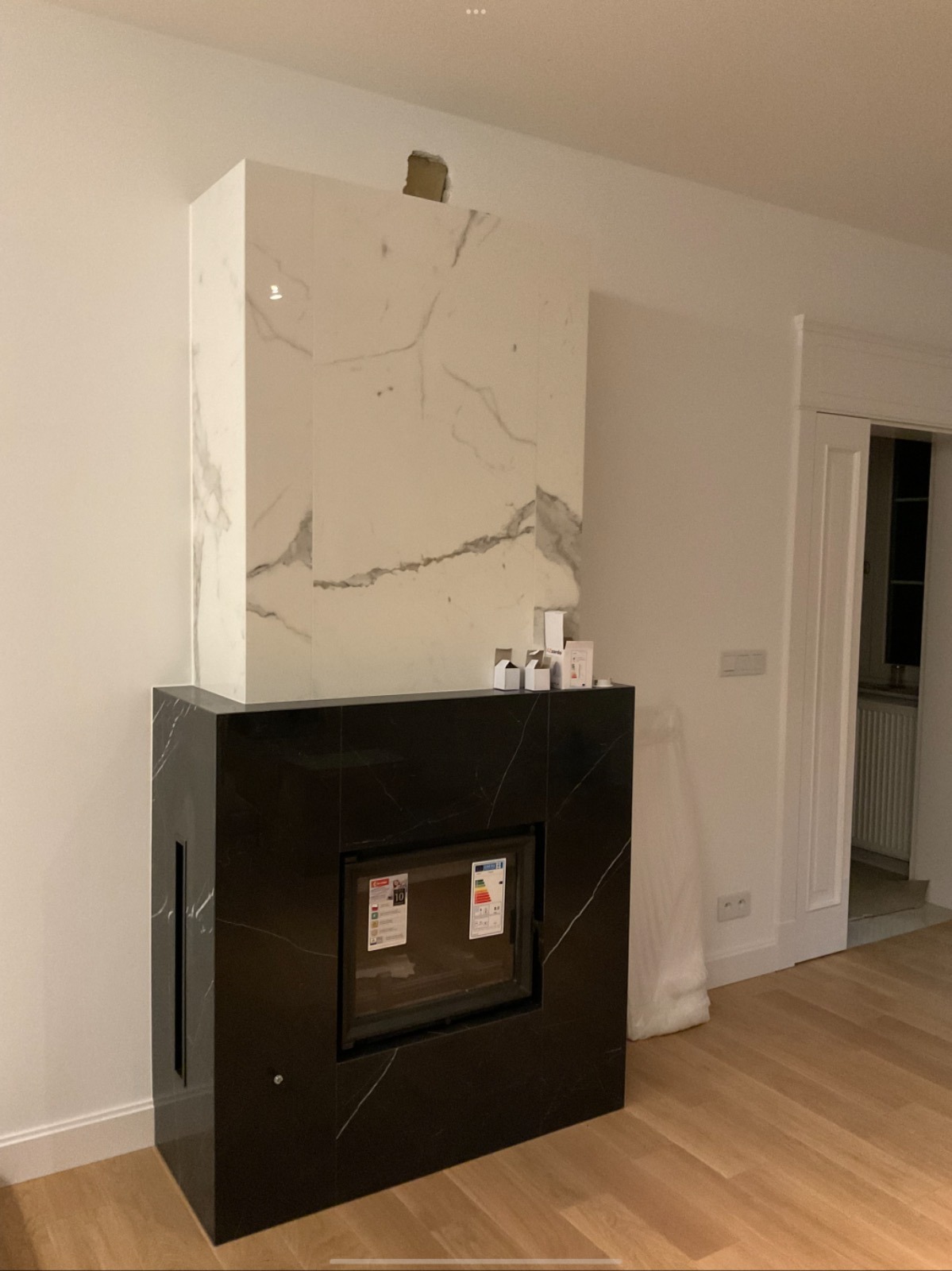 Nowoczesny kominek z czarnym marmurem na dole i białym marmurem na górze, widoczny otwór w ścianie nad kominkiem, podłoga z jasnego drewna.