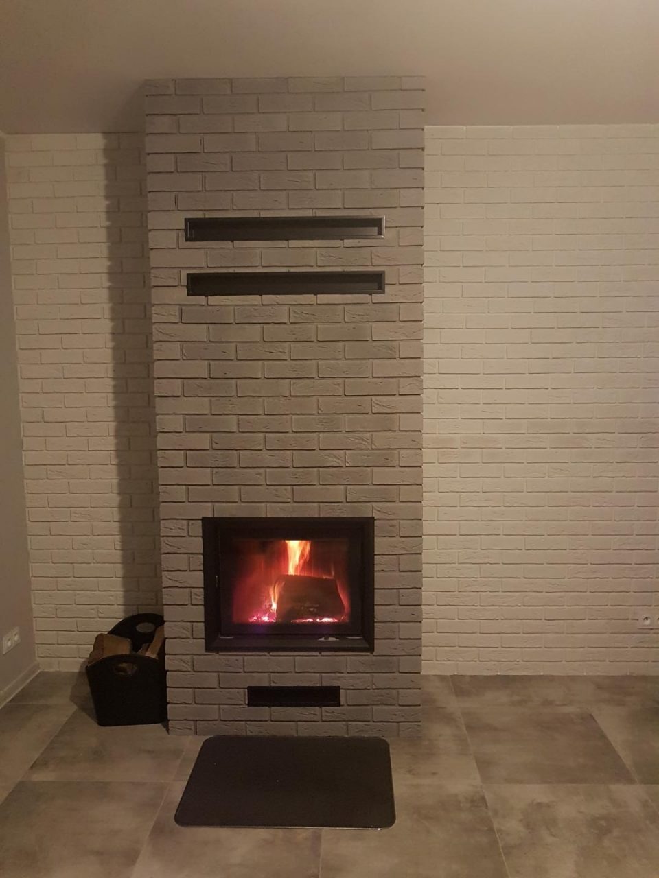 Nowoczesny kominek z obudową z szarej cegły, z czarnym wkładem kominkowym i płomieniami, w tle ściana z białej cegły i szara podłoga z płytek.