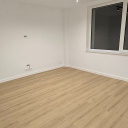 Instalpos - Puste, świeżo wykończone wnętrze z panelami podłogowymi w odcieniach beżu, białymi ścianami z widocznymi gniazdkami elektrycznymi i otworem na kabel, oraz dużym oknem z białymi ramami.