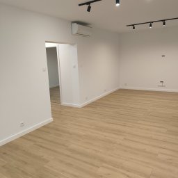 Instalpos - Puste, świeżo wykończone wnętrze z jasną podłogą imitującą drewno, białymi ścianami, listwami przypodłogowymi i oświetleniem szynowym na suficie. Widoczny klimatyzator i gniazdka elektryczne.