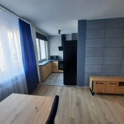 Instalpos - Nowoczesne wnętrze: drewniana podłoga, szara ściana z płytkami, drewniana szafka RTV, czarna lodówka, kuchnia z drewnianymi frontami i oknem z niebieską zasłoną.