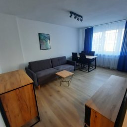 Instalpos - Salon z szarą sofą, stolikiem kawowym z metalową podstawą, drewnianą komodą i szafką RTV, oraz stołem z krzesłami w tle przy oknie z niebieskimi zasłonami.