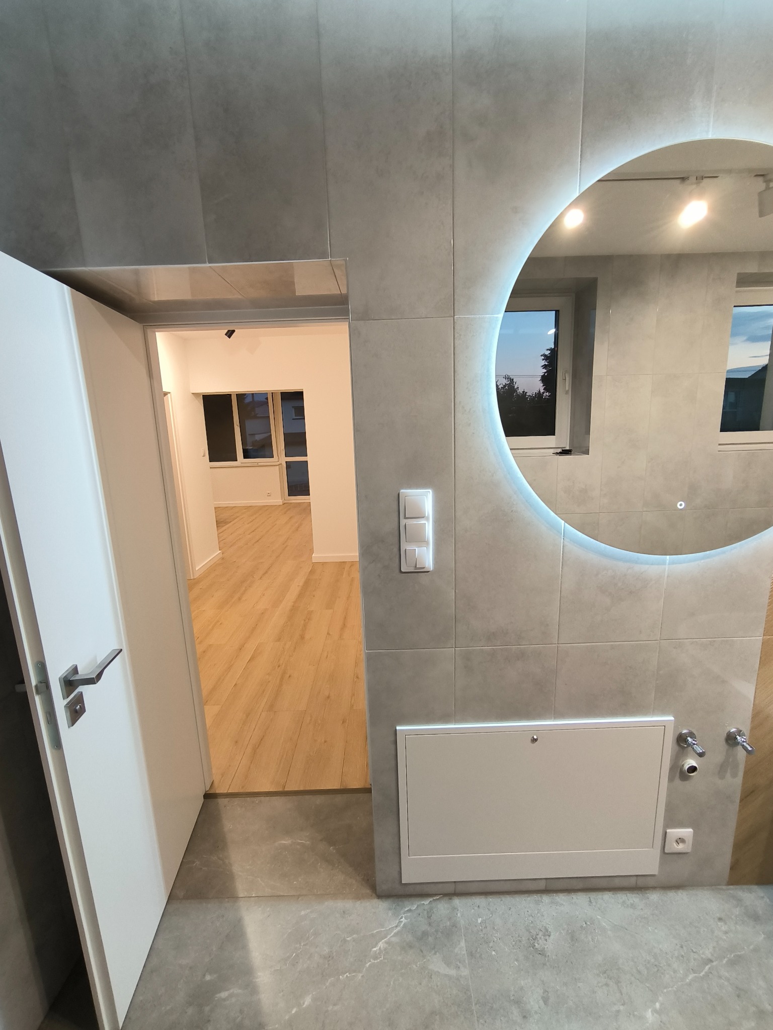 Wykończona łazienka z szarą glazurą, okrągłym lustrem z podświetleniem LED i widokiem na nowo wykończony pokój z drewnianą podłogą przez otwarte drzwi.