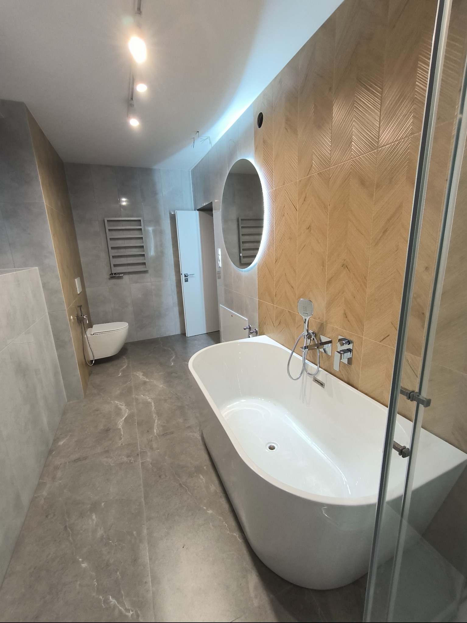 Nowoczesna łazienka z wolnostojącą wanną, okrągłym lustrem z podświetleniem LED i ścianą wyłożoną płytkami w jodełkę, szara podłoga z efektem marmuru.