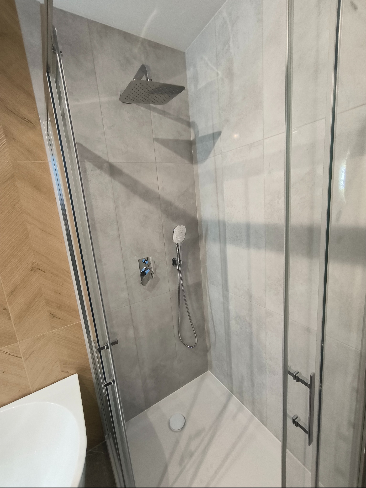 Nowoczesna kabina prysznicowa z deszczownicą i baterią termostatyczną, wykończona szarymi płytkami imitującymi kamień, widok z boku na szklane drzwi i fragment umywalki.