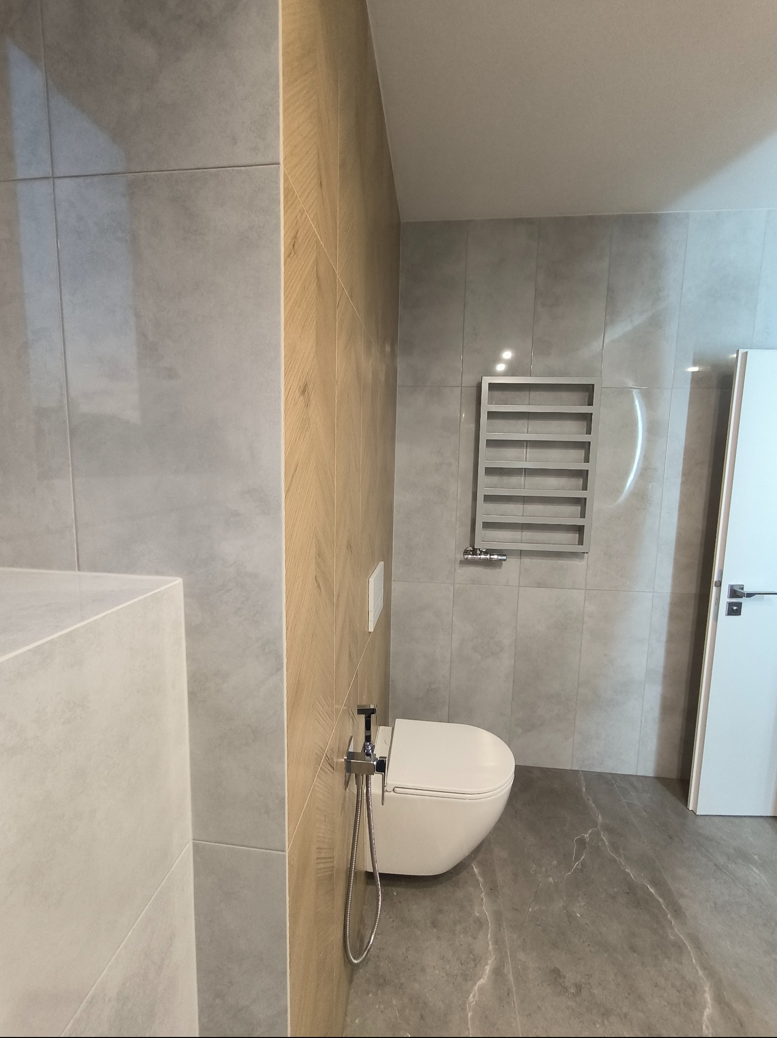 Nowoczesna łazienka z szarymi płytkami imitującymi marmur na podłodze, drewnopodobną ścianą, wiszącą toaletą z bidetką i grzejnikiem drabinkowym.