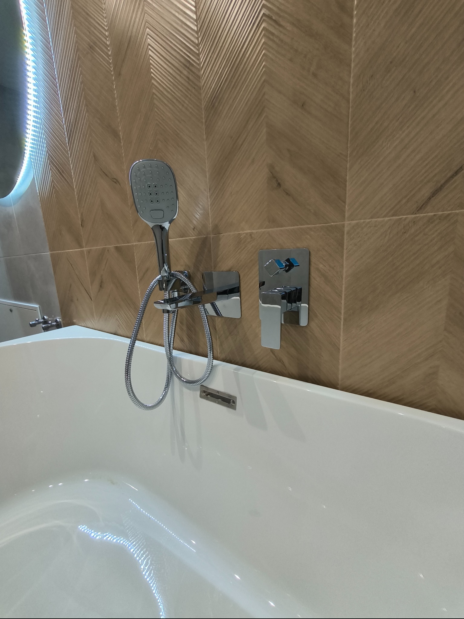 Biała wanna z chromowaną baterią, słuchawką prysznicową i brązowymi płytkami ułożonymi w jodełkę na ścianie.