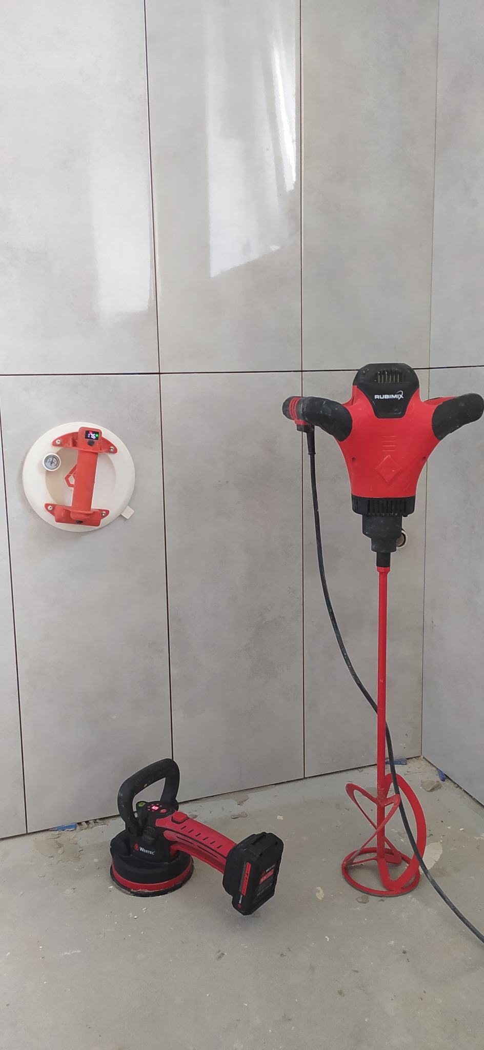 Nierozpoczęte układanie płytek ściennych w łazience: widoczna przyssawka próżniowa do płytek na ścianie, mieszadło do zaprawy i przyssawka akumulatorowa do przenoszenia płytek na podłodze.