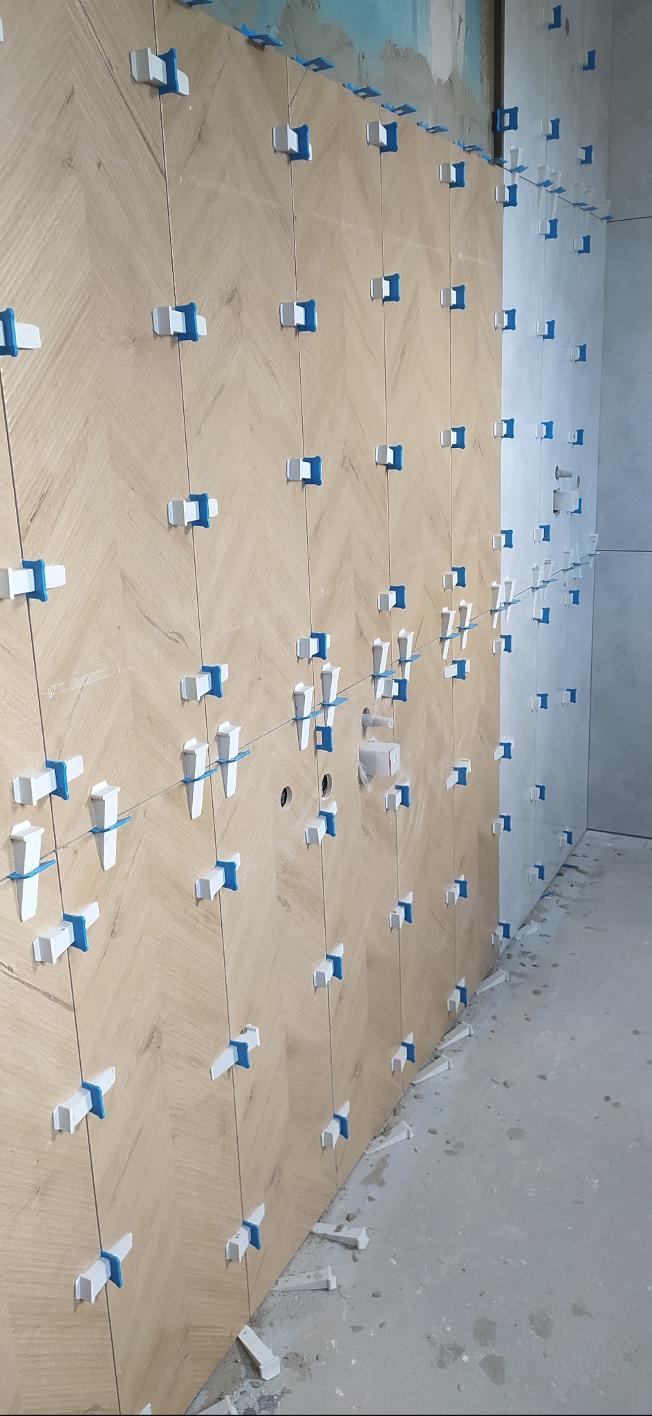 Układanie płytek imitujących drewno w jodełkę na ścianie, widoczne niebiesko-białe klipsy poziomujące i sznurki, surowy stan wykończenia pomieszczenia.
