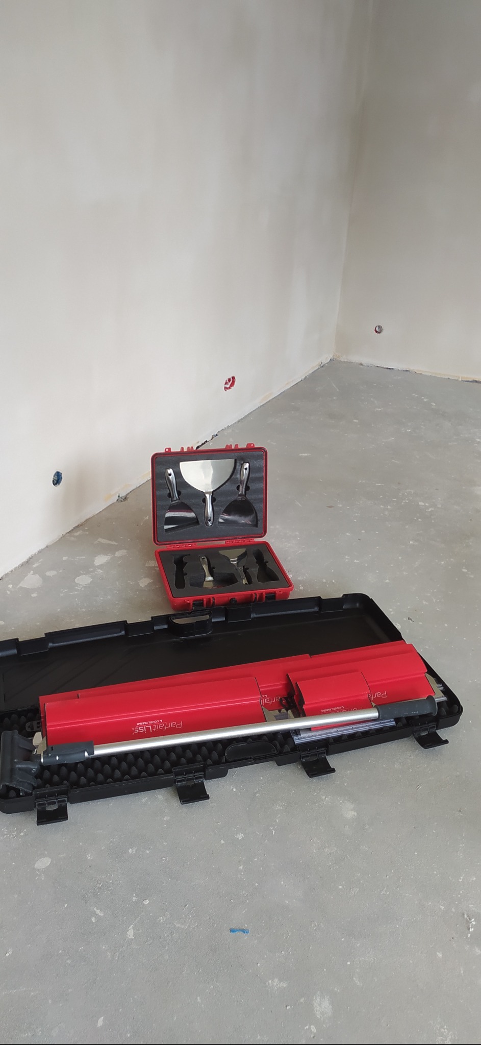 Zestaw narzędzi szpachlarskich ParfaitLiss w czerwonej i czarnej walizce na tle surowych ścian i podłogi w trakcie prac wykończeniowych.