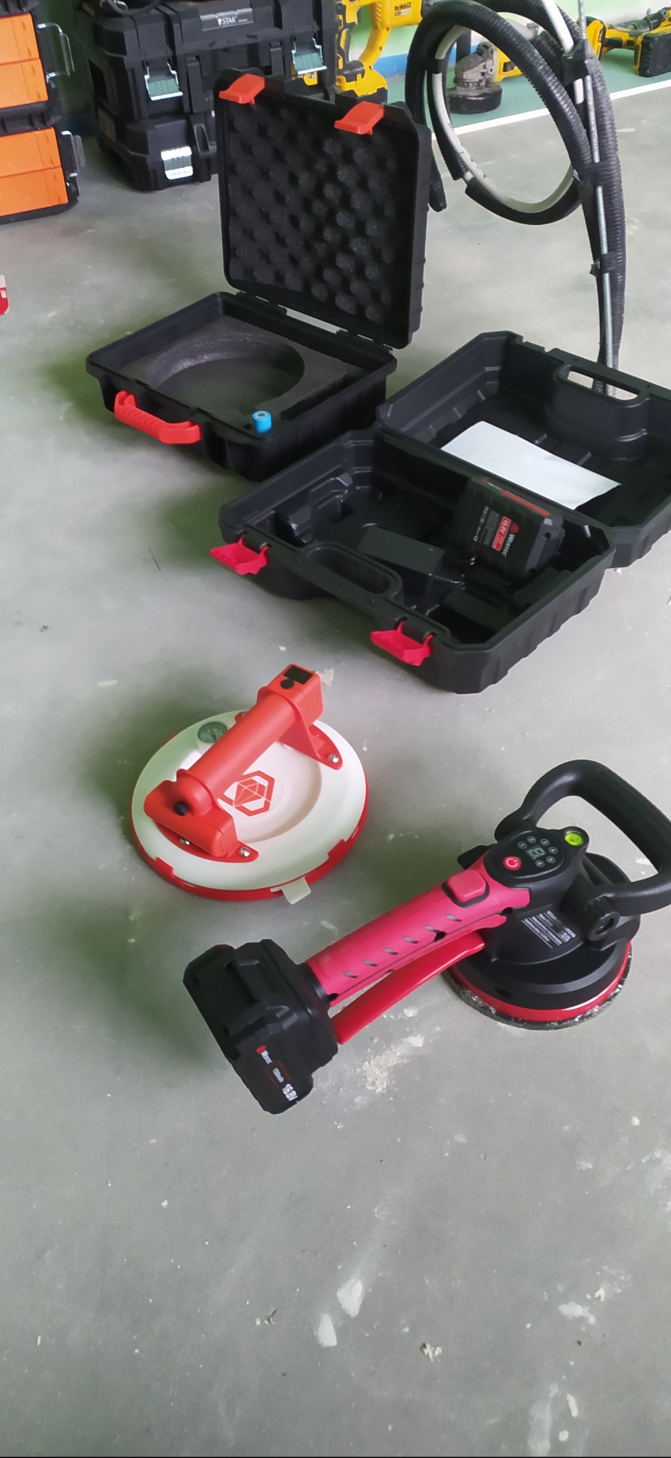 Dwa akumulatorowe uchwyty do przenoszenia płytek, jeden czerwono-biały z logo Rubiego, drugi czarno-różowy, obok otwartych czarnych skrzynek narzędziowych na szarym tle podłogi.