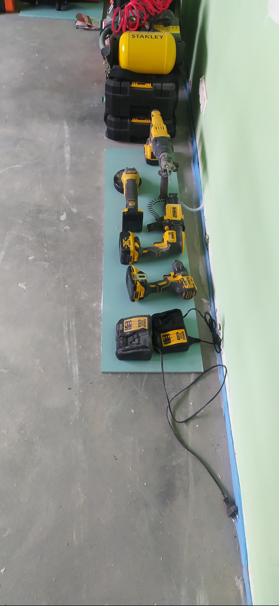 Zestaw narzędzi DeWalt: wiertarka udarowa, szlifierka kątowa, wkrętarka, zakrętarka i ładowarki akumulatorów ułożone na płycie gipsowej, obok stoi kompresor Stanley.