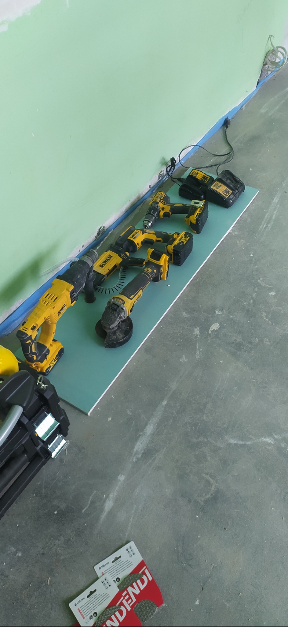 Różnorodne elektronarzędzia marki DeWalt ułożone na płycie gipsowo-kartonowej przy zielonej ścianie, widoczne wiertarki, szlifierka kątowa i piła szablasta, obok opakowania tarcz diamentowych...