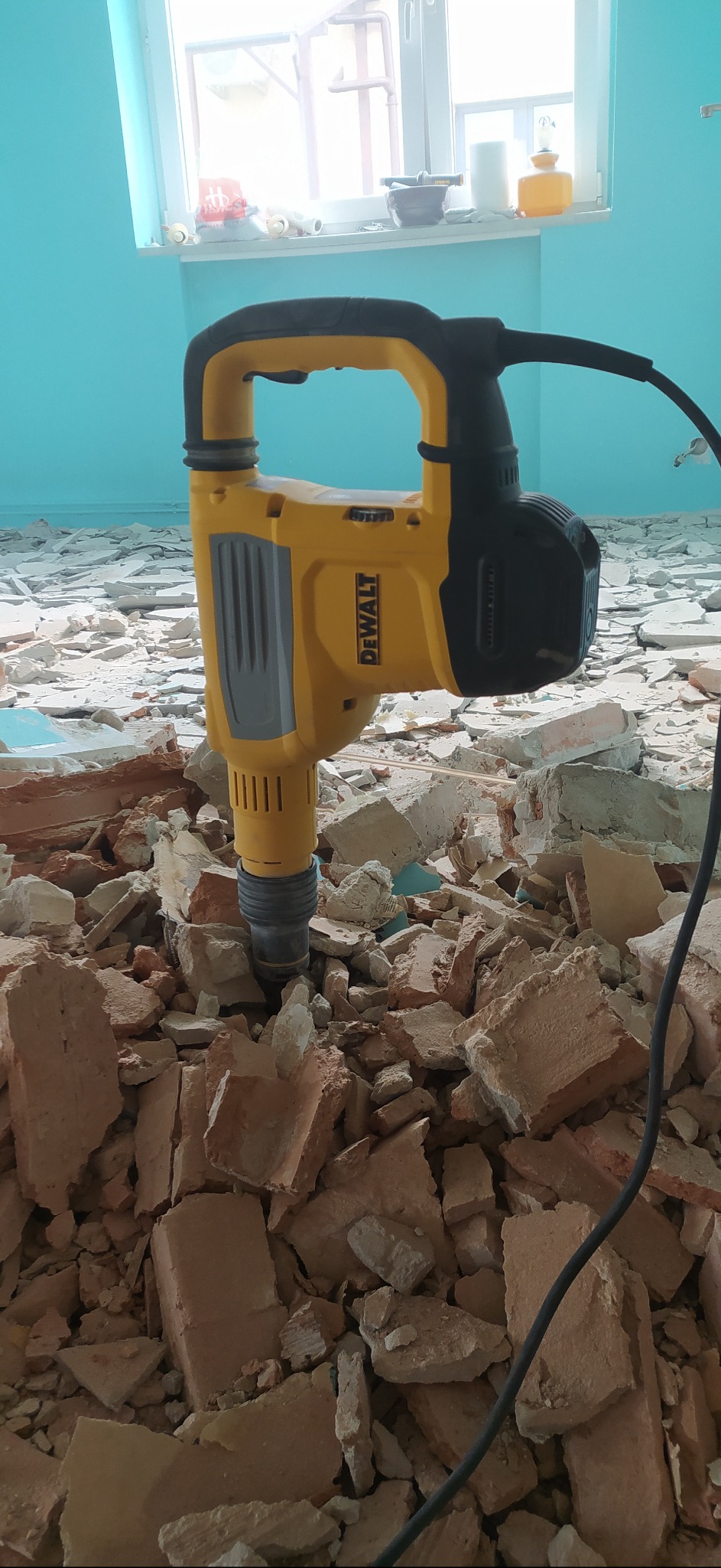 Żółto-czarny młot udarowy DeWalt na stercie gruzu ceglanego w pomieszczeniu z błękitnymi ścianami, widoczne okno w tle.