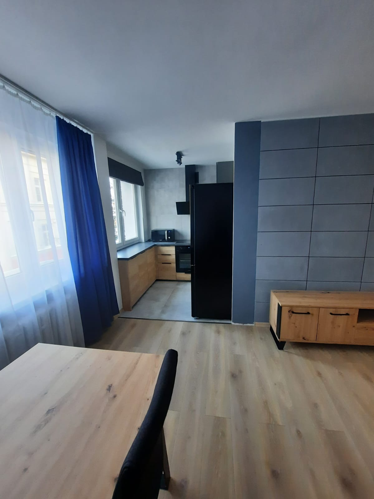 Nowoczesne wnętrze: drewniana podłoga, szara ściana z płytkami, drewniana szafka RTV, czarna lodówka, kuchnia z drewnianymi frontami i oknem z niebieską zasłoną.