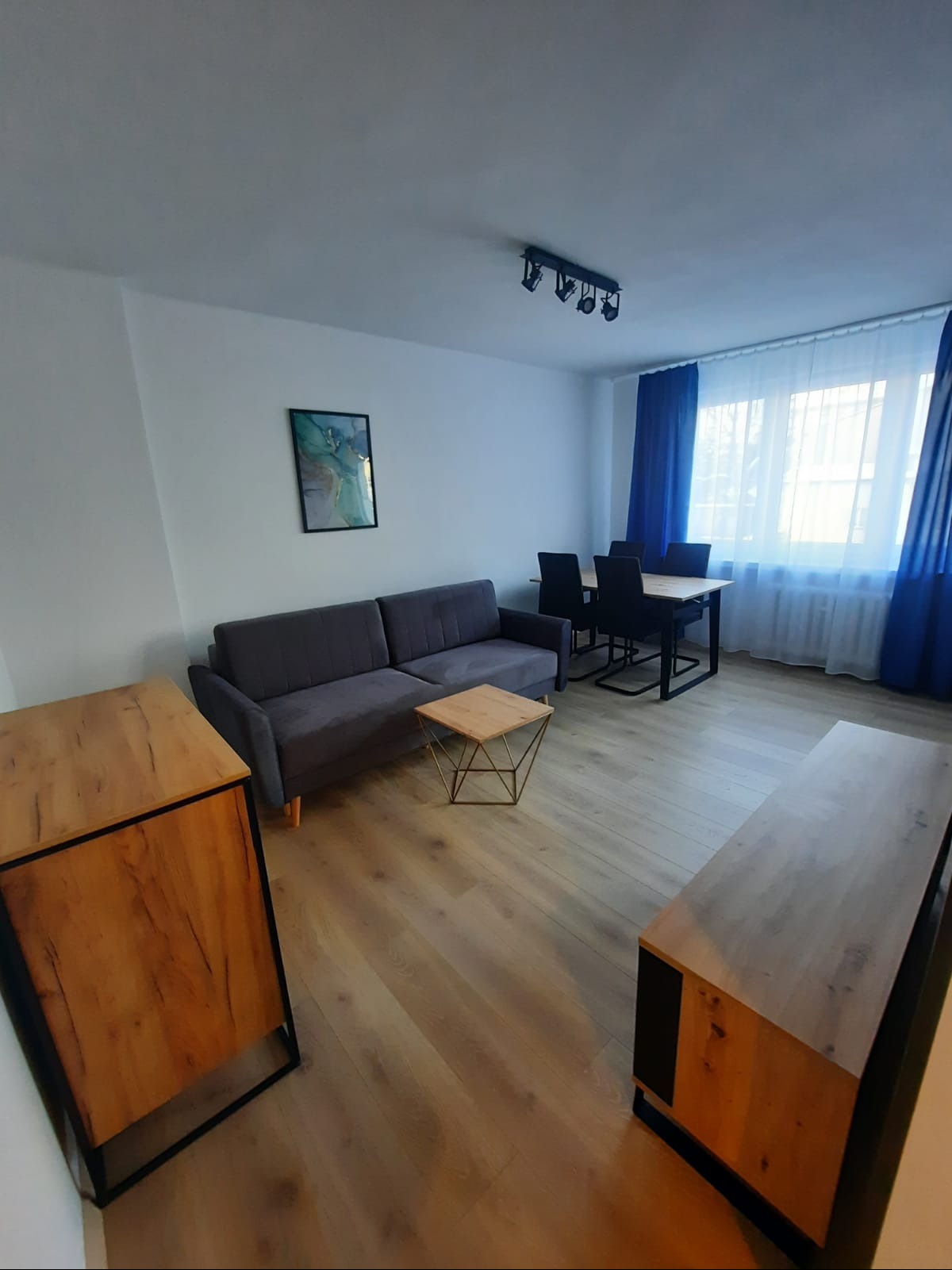 Salon z szarą sofą, stolikiem kawowym z metalową podstawą, drewnianą komodą i szafką RTV, oraz stołem z krzesłami w tle przy oknie z niebieskimi zasłonami.