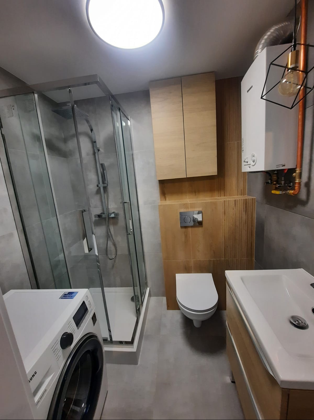 Nowoczesna łazienka z kabiną prysznicową, pralką, umywalką z szafką, toaletą podwieszaną i grzejnikiem gazowym, wykończona w szarościach i drewnie.